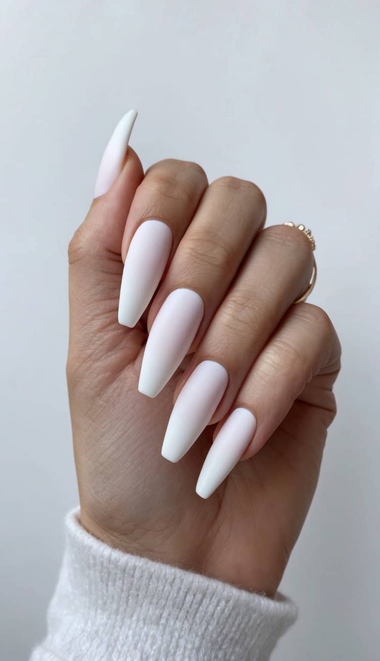 Hand raised against a neutral wall showing matte wedding nails in pure white with long tapered coffin silhouettes and a subtle gold band on the finger.