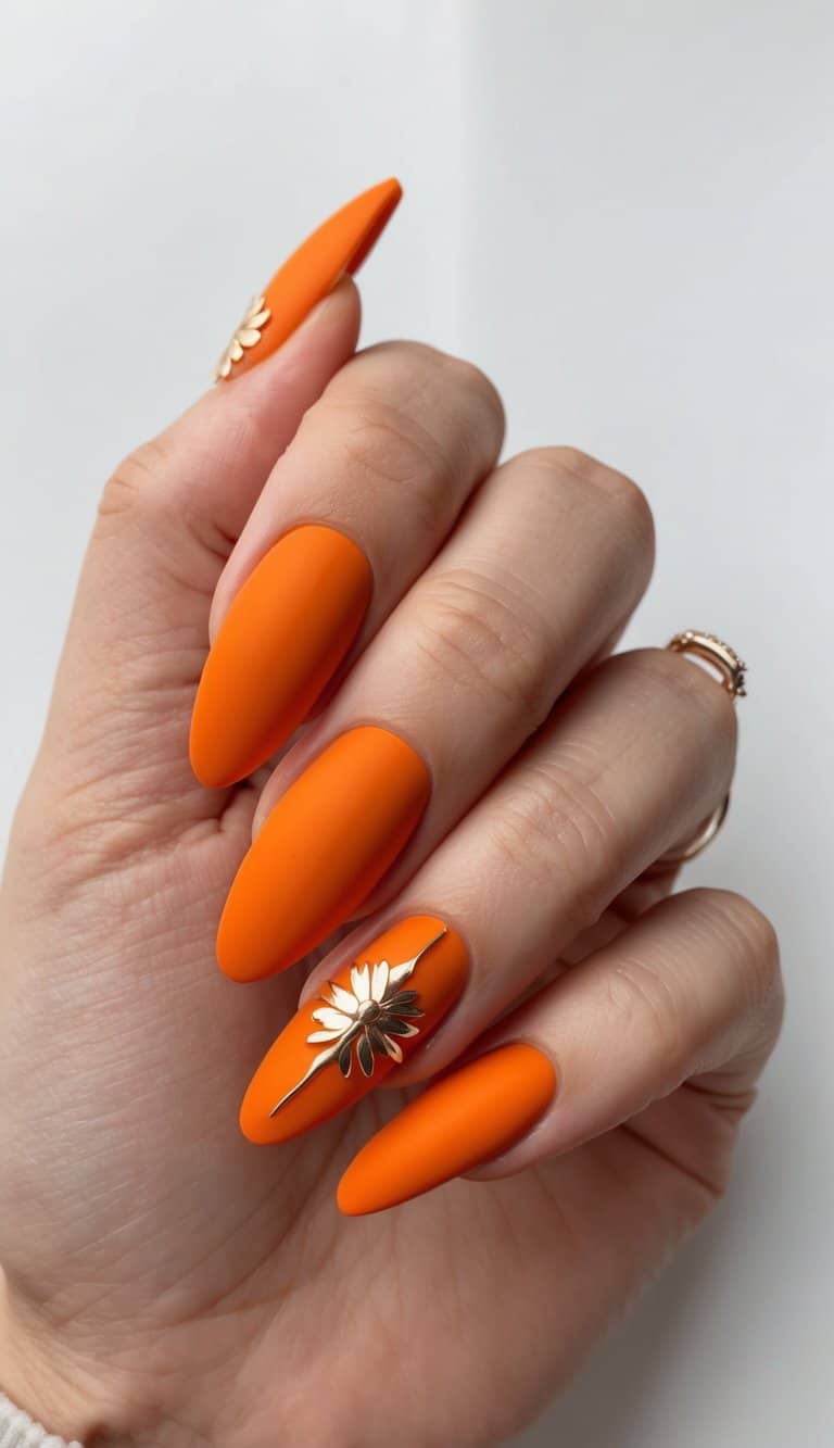 Hand posed upright showcasing matte wedding nails in bright sunset orange, almond-shaped with a raised gold flower design on one accent nail.