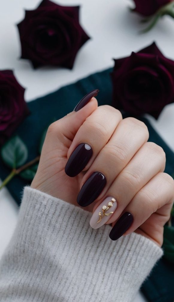 A hand with dark plum wedding nails in a glossy finish, paired with a matte nude accent nail decorated with elegant gold floral embellishments and tiny pearl-like studs.