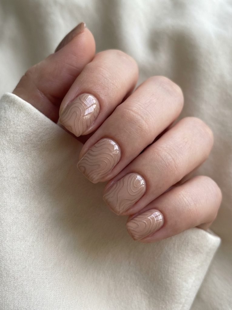 Close-up of a hand wrapped in cream fabric featuring short square wedding nails in warm beige nude with glossy raised taupe swirl patterns on each nail.