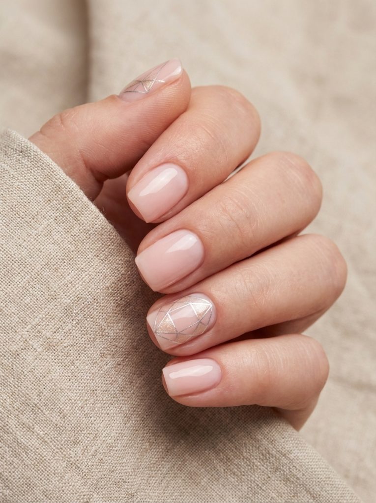 Close-up of a hand holding beige fabric, showcasing short square wedding nails in glossy blush nude with one accent nail featuring fine silver geometric lines over a soft white shimmer base.