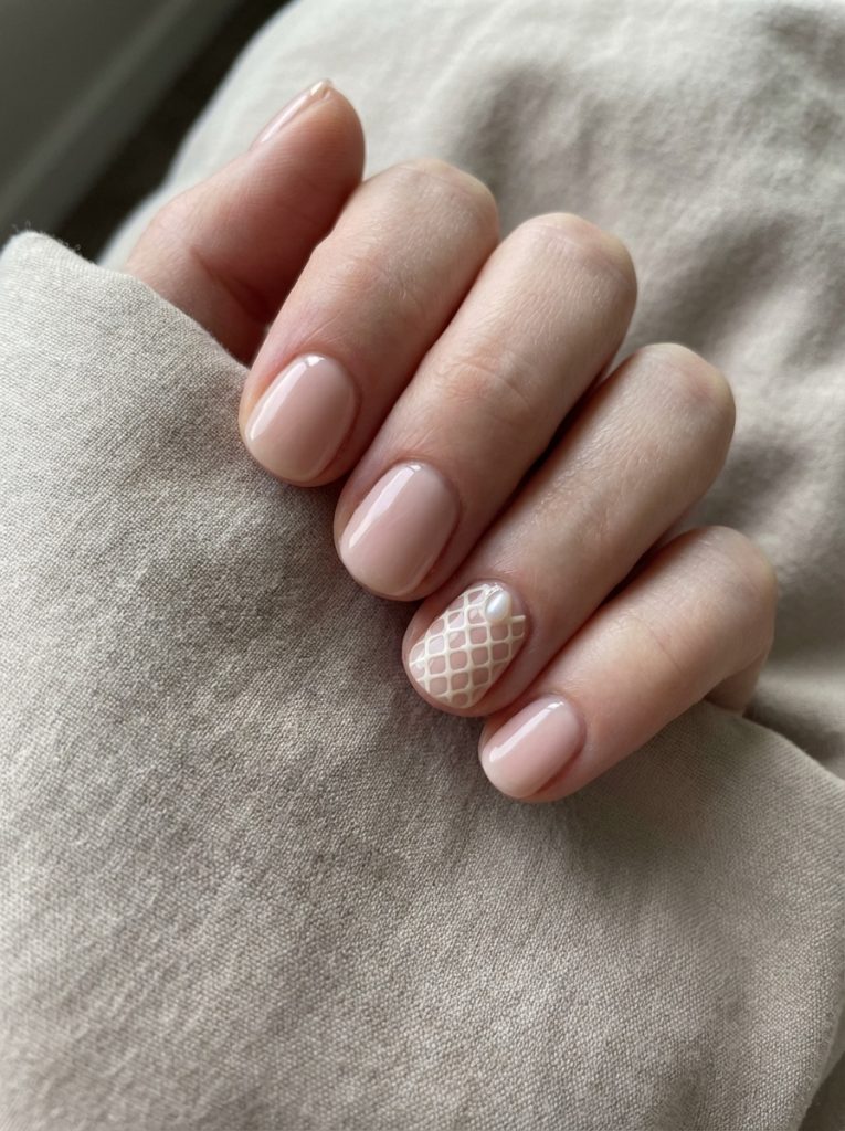 A softly posed hand against neutral fabric featuring short wedding nails in glossy blush nude, with one nail detailed in a delicate white lattice pattern over sheer pink.