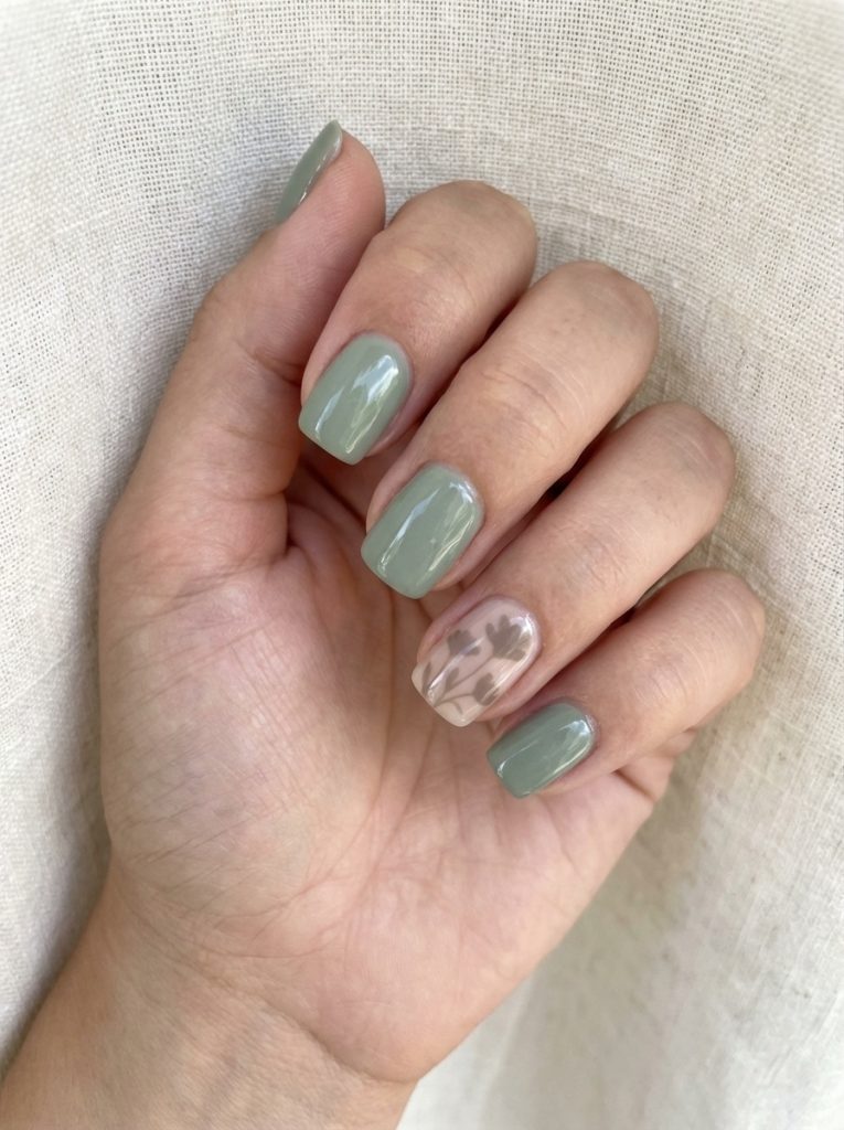 Softly lit hand against linen background featuring short square wedding nails in dusty sage green and one milky pink accent nail with subtle botanical leaf design.