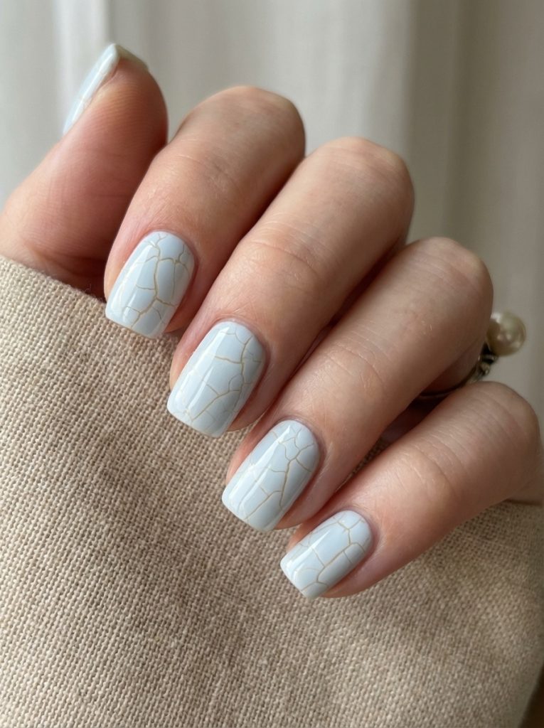 A close view of a hand wrapped in beige knit fabric featuring short square wedding nails in pale porcelain blue, detailed with fine gold crackle lines across each glossy nail.