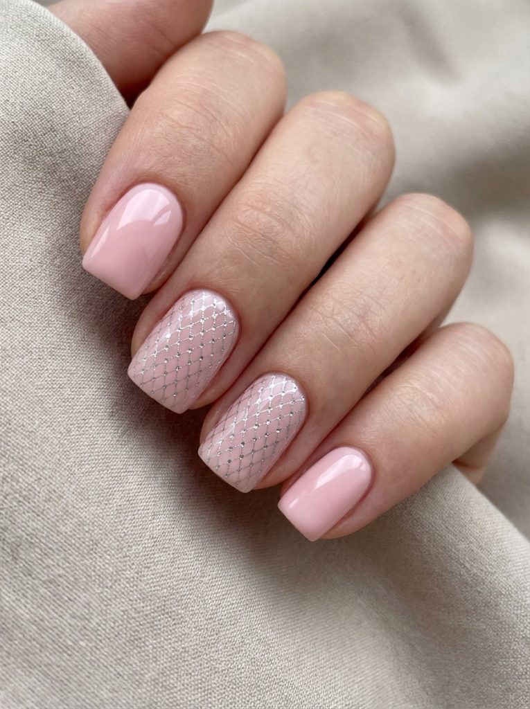 A close-up of a hand resting on beige fabric featuring short square wedding nails in glossy blush pink, with two accent nails designed in nude polish and delicate silver lattice detailing.
