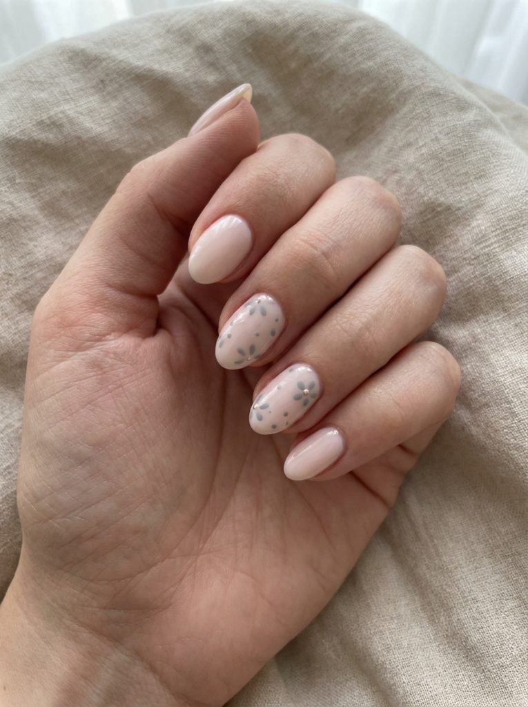 Hand resting on textured beige fabric, featuring short wedding nails in sheer blush polish with two oval accent nails decorated in small grey-blue floral dots.