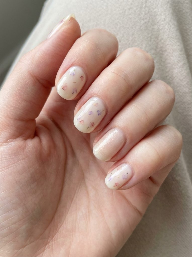 Manicured fingers curled softly, displaying short wedding nails with translucent ivory polish, scattered lilac and blush floral flecks, and one fine glitter accent nail.