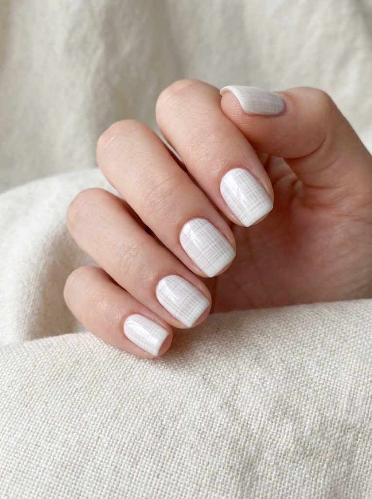 A close-up of a hand resting on cream fabric showcasing short square wedding nails in soft white polish detailed with a fine crosshatch grid pattern and glossy shine.