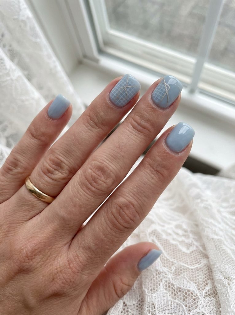 Close-up of a hand wearing a gold band, highlighting short square wedding nails in dusty baby blue with delicate white grid accents on selected nails.