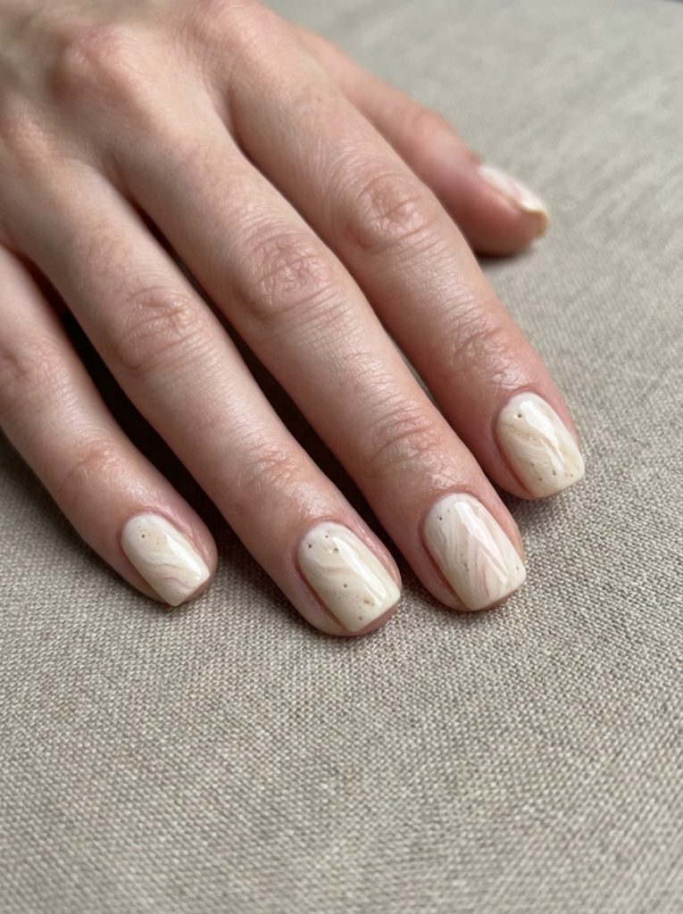 A hand resting on textured beige fabric showcases short square wedding nails in creamy oatmeal nude with soft marble swirls and delicate brown speckled detailing.