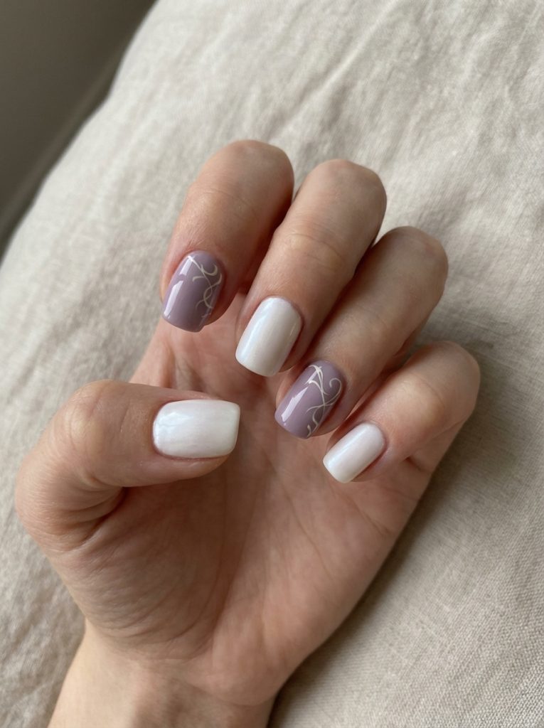 Hand resting on textured linen, showcasing square short wedding nails in white and soft lavender with elegant ivory swirl detailing on accent nails.