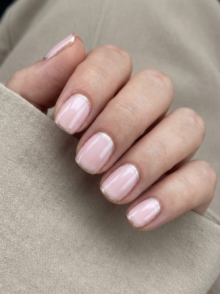 A hand wrapped in beige fabric showcasing short square wedding nails in glossy blush pink, each outlined at the cuticle with fine champagne glitter detailing.