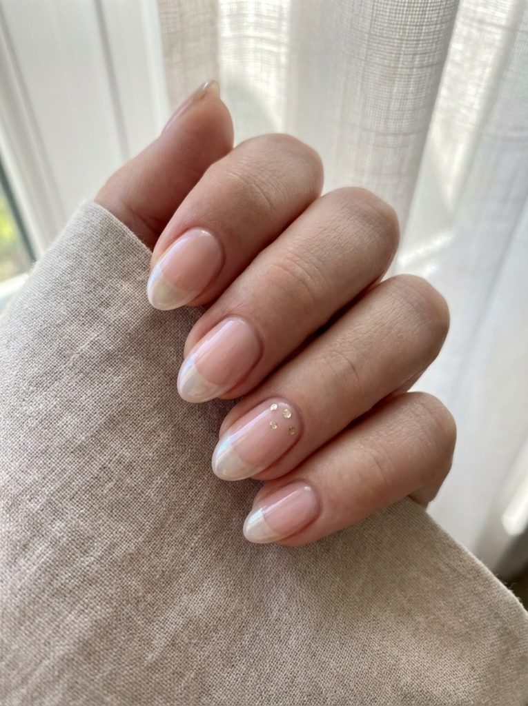 Close view of a manicured hand resting on linen, featuring short wedding nails in pink-beige polish with delicate French tips and tiny gold studs arranged on one accent nail.