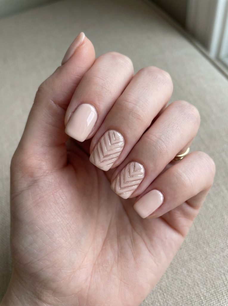 A hand near a window featuring short square wedding nails in creamy blush nude, with two accent nails detailed in raised chevron patterns and subtle champagne shimmer.