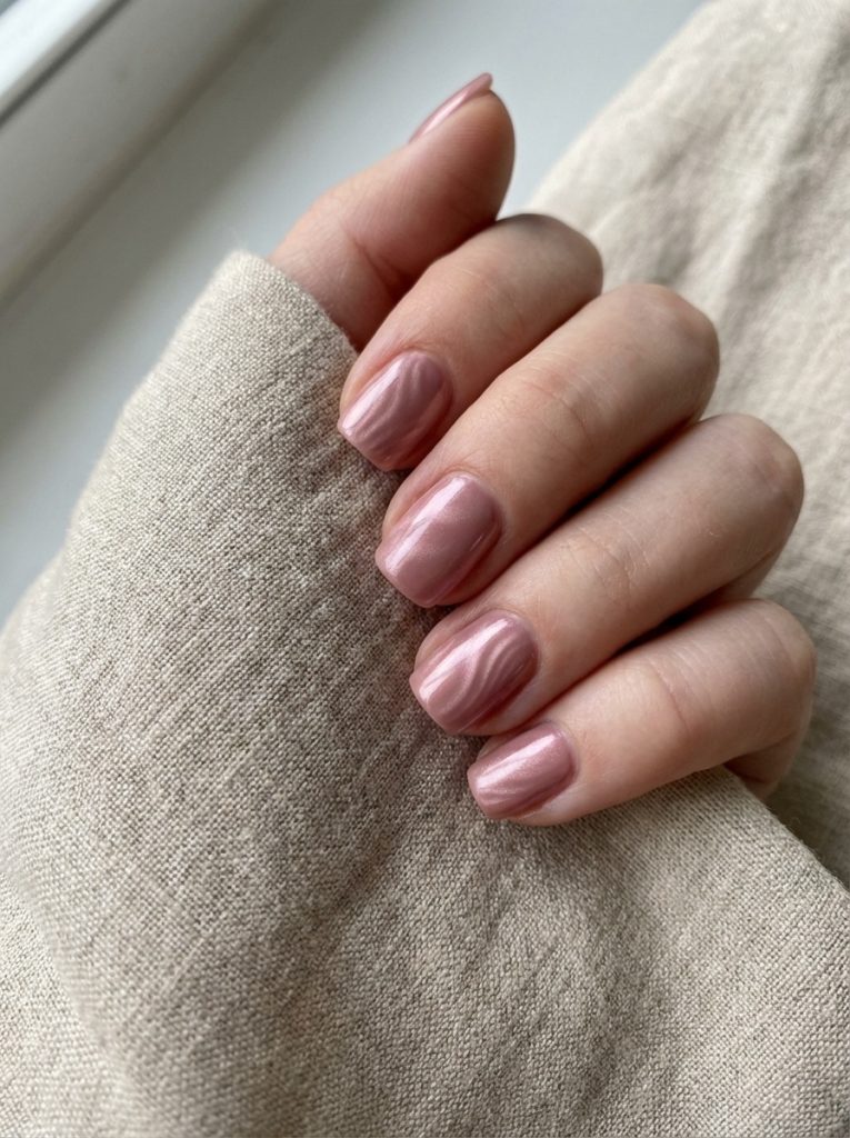 A hand wrapped in beige linen showcasing short square wedding nails in rosy mauve chrome polish with a smooth pearlescent, reflective finish.