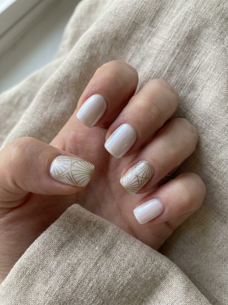 A close-up of a hand resting on beige linen showcasing short square wedding nails in pearl white, with accent nails detailed in delicate gold fan-inspired line patterns.
