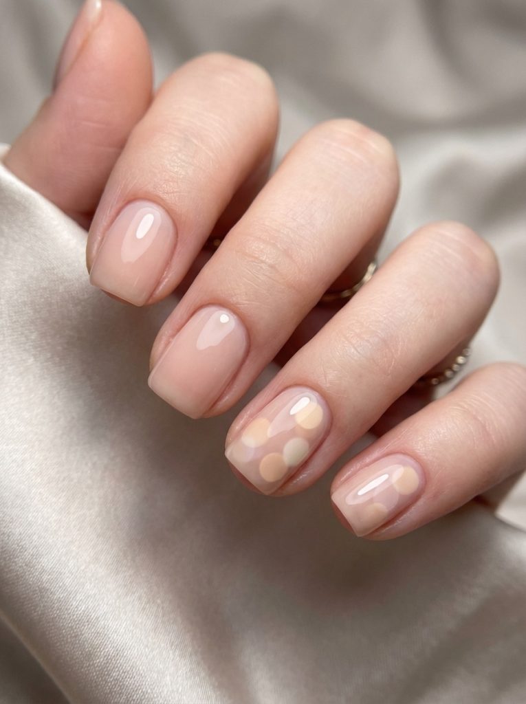 A close-up of a hand wrapped in satin fabric showcasing short square wedding nails in sheer blush nude, with two accent nails decorated in soft cream polka dots.