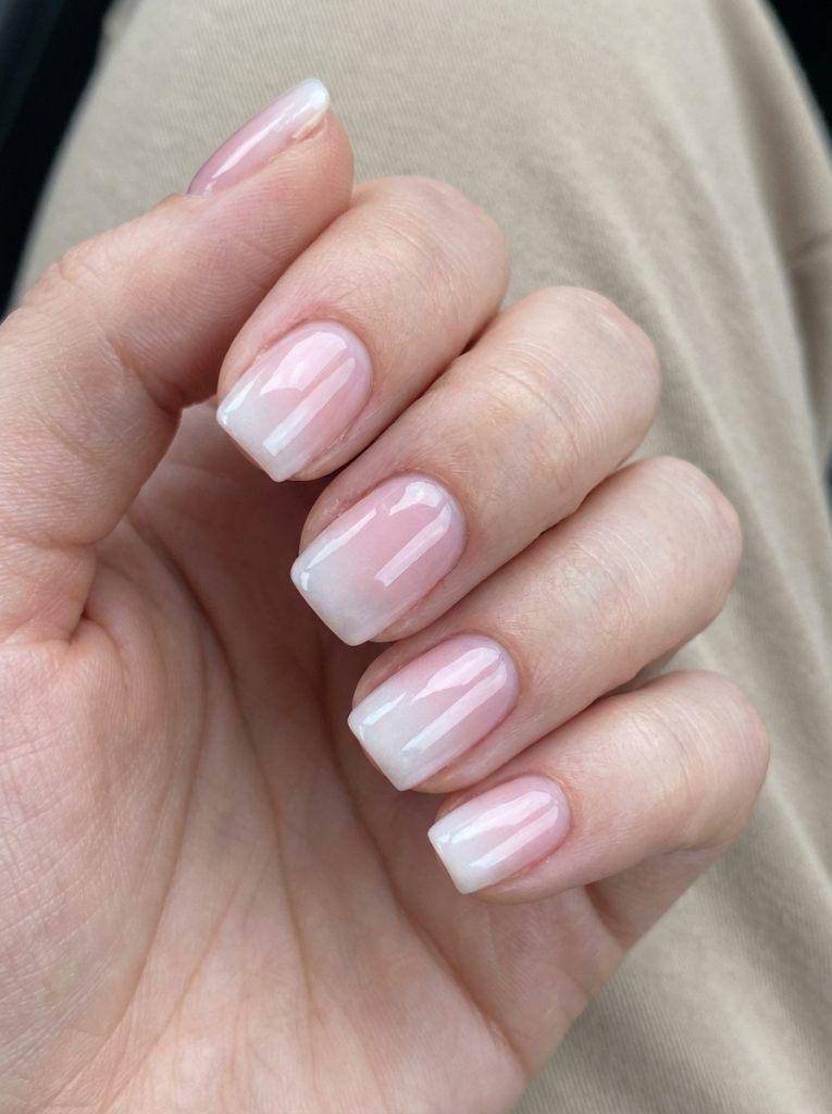 Fingers gently curled over neutral fabric display short square wedding nails in a blush-to-white ombré design with a smooth glossy surface.