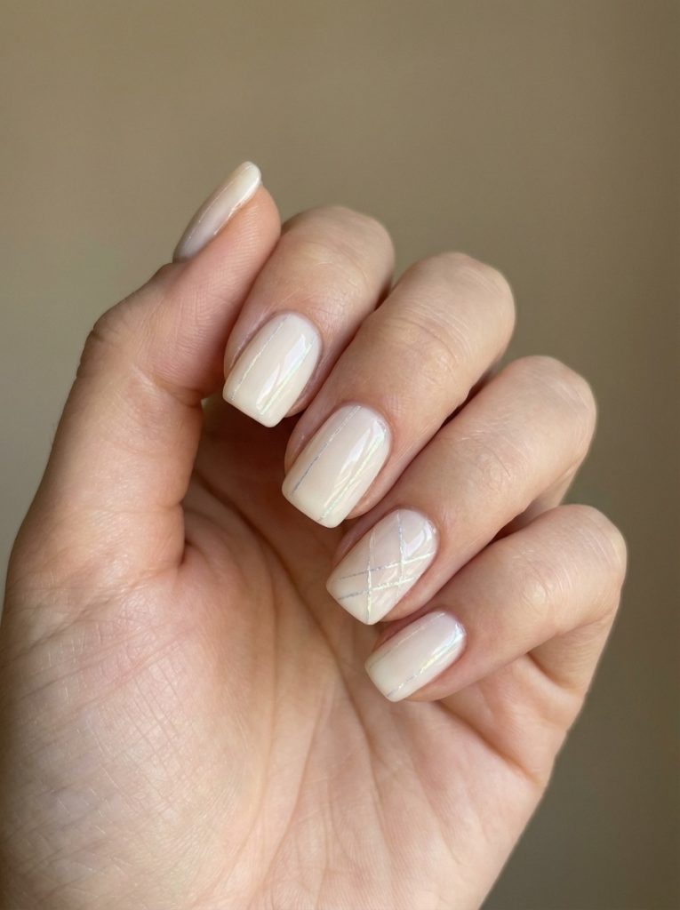 Natural hand posed softly, displaying short wedding nails in milky taupe with one accent nail showcasing subtle crisscross line design in pale metallic tones.