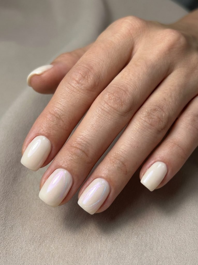Hand gently placed on beige textile, displaying square short wedding nails in creamy beige polish with a soft pearly shimmer.