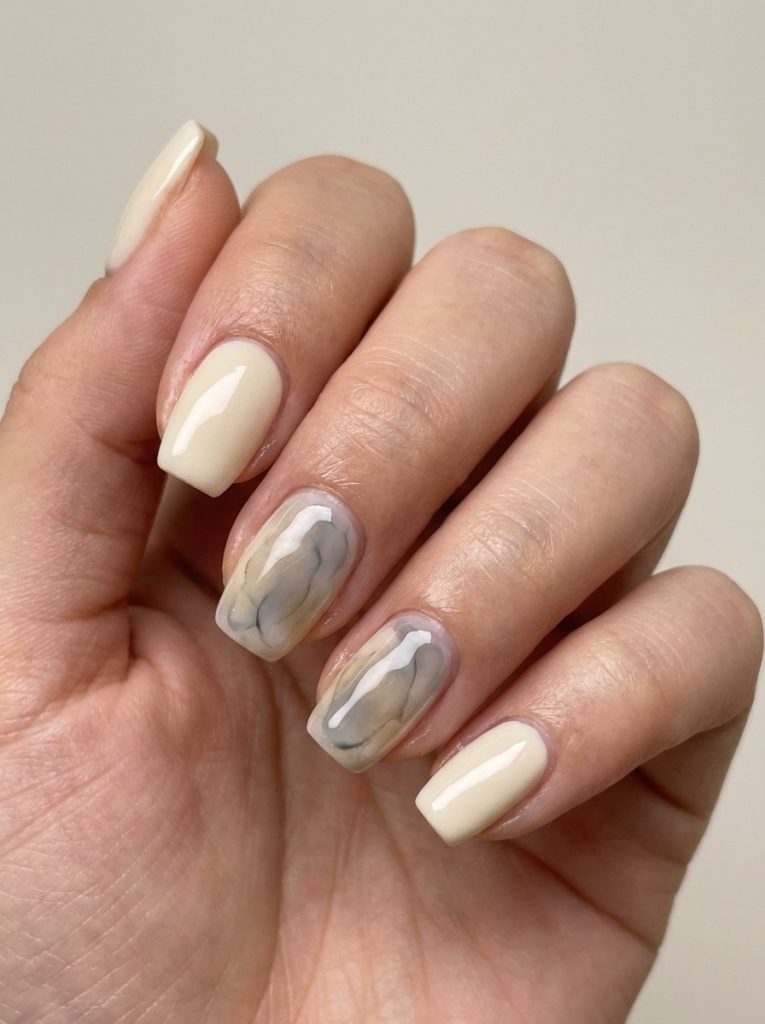 Close-up of a manicured hand showing short wedding nails in ivory-beige polish with two nails designed in soft grey marble patterns.