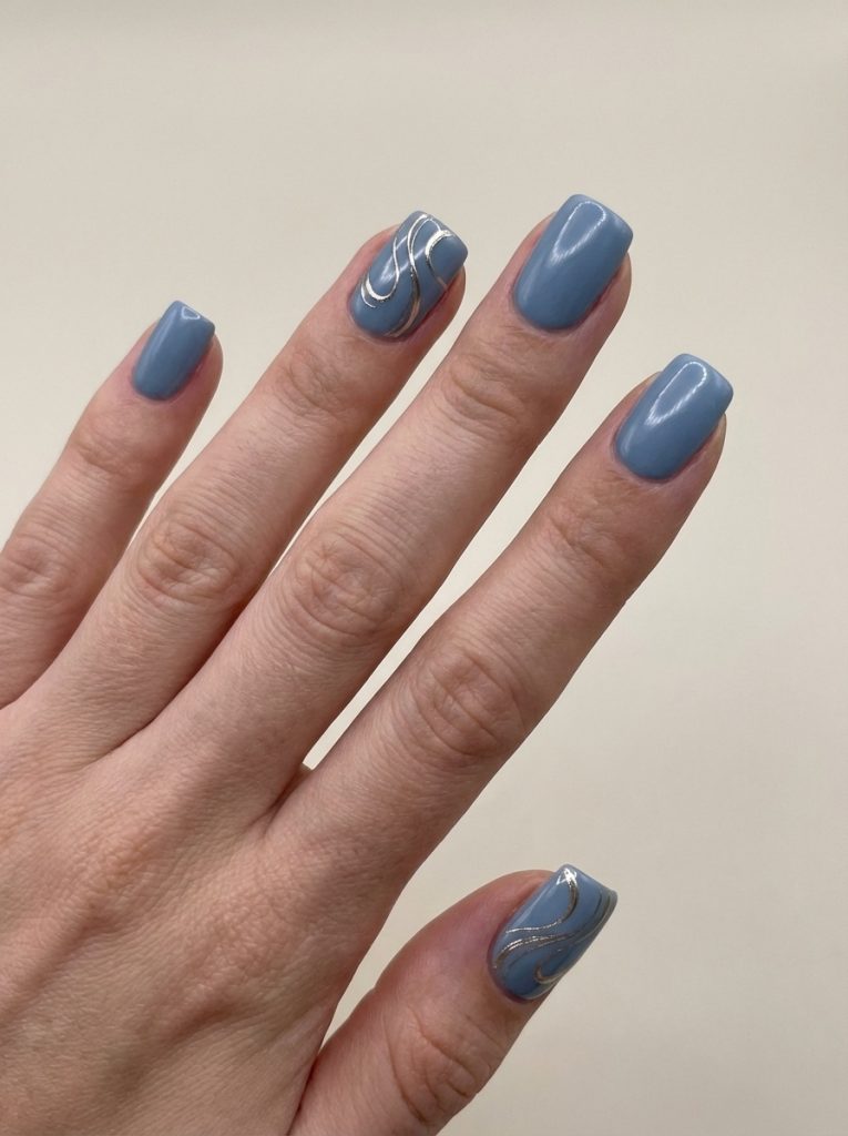 Fingers gently spread showcase short square wedding nails painted in dusty denim blue, highlighted by accent nails with fine gold ribbon-style swirl detailing.