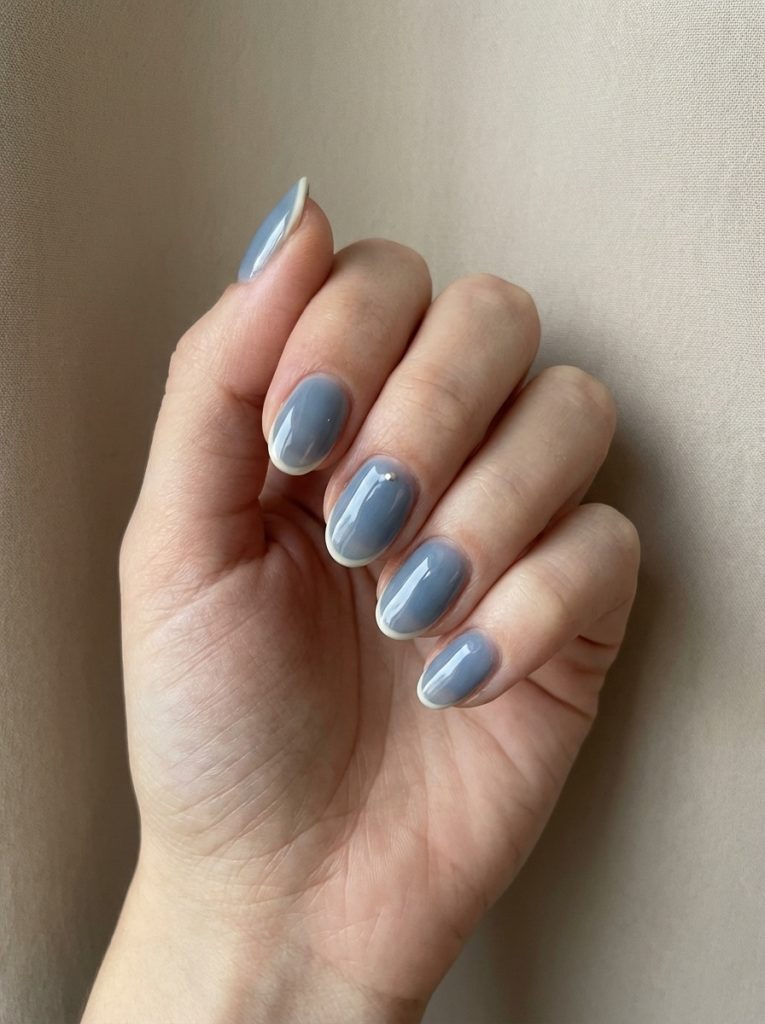 Natural hand shown up close, featuring short wedding nails in steel-blue polish accented with thin creamy French tip borders.