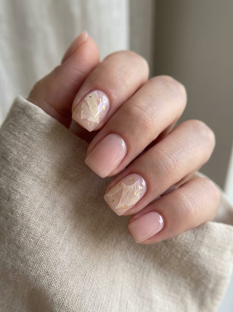 Close view of a hand highlighting short square wedding nails in creamy blush polish, complemented by translucent accent nails decorated with soft leaf designs and gold flecks.
