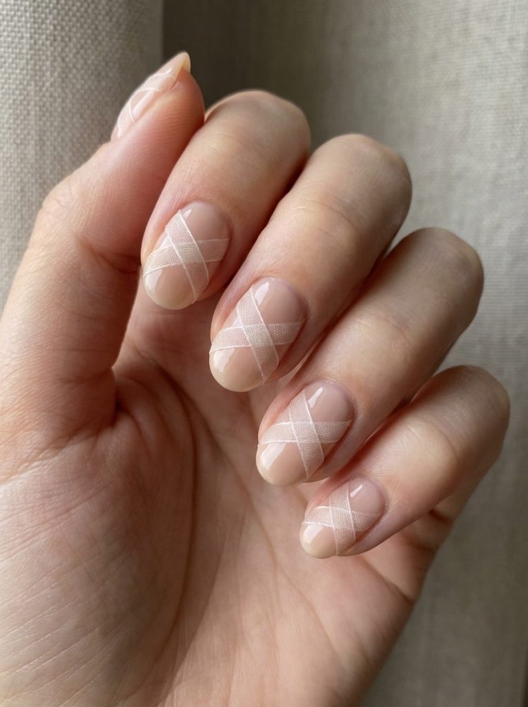 A softly lit hand displays nude wedding nails with a translucent beige-pink base, accented by fine crisscross lines that resemble delicate ribbon layered over glossy nails.