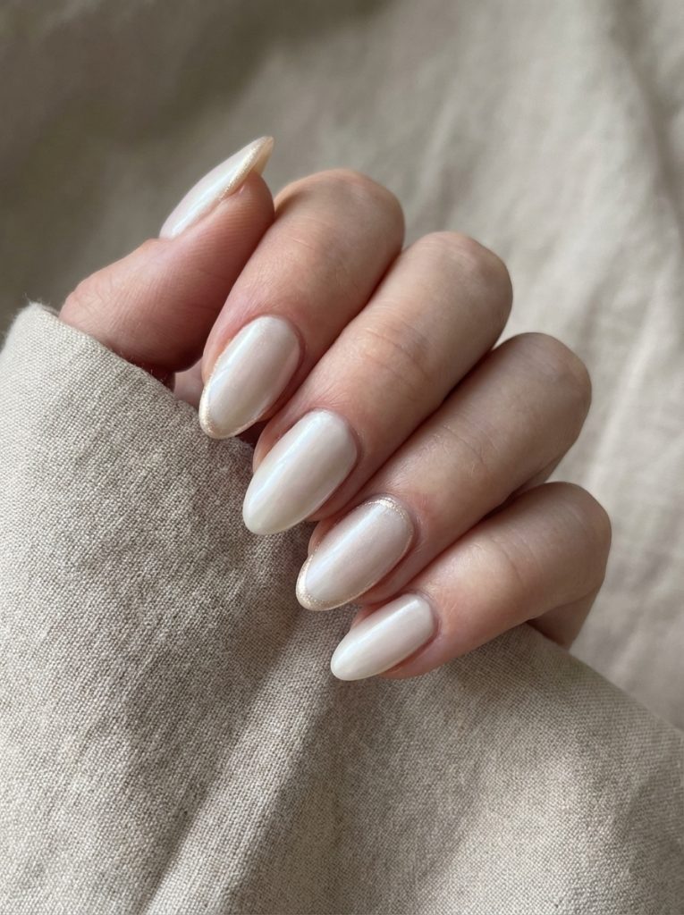 Close-up of a hand holding textured beige fabric, showcasing pearl wedding nails in creamy champagne ivory with delicate shimmer outlining the almond-shaped tips.