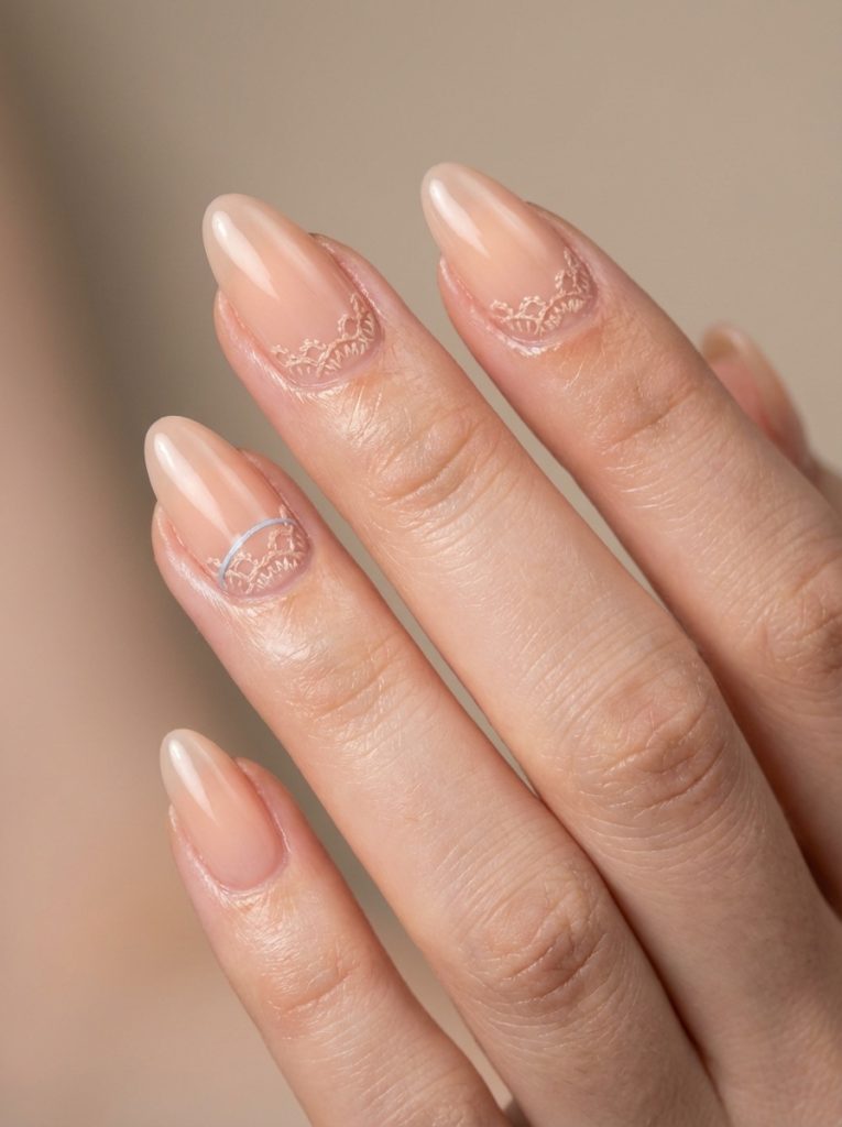 Close-up of a hand showing nude wedding nails in a translucent peach tone, featuring fine lace-style detailing along the cuticle line and a smooth glossy finish.