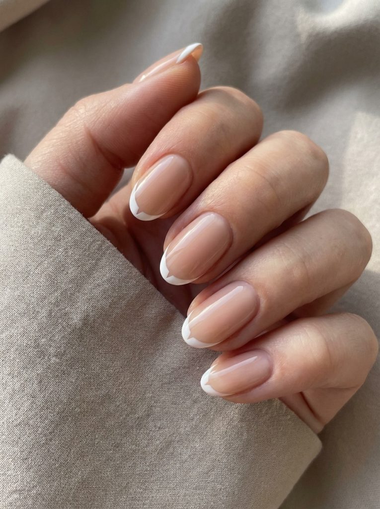 Hand posed against a beige textile background featuring nude wedding nails with a glossy blush-toned base and thin white French tips shaped neatly at the nail ends.