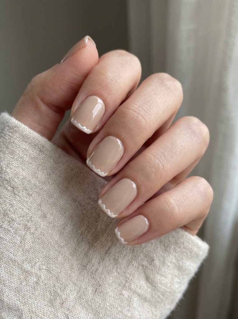 Manicured hand photographed in soft daylight, displaying nude wedding nails with a creamy beige gloss and gently scalloped white tip accents.