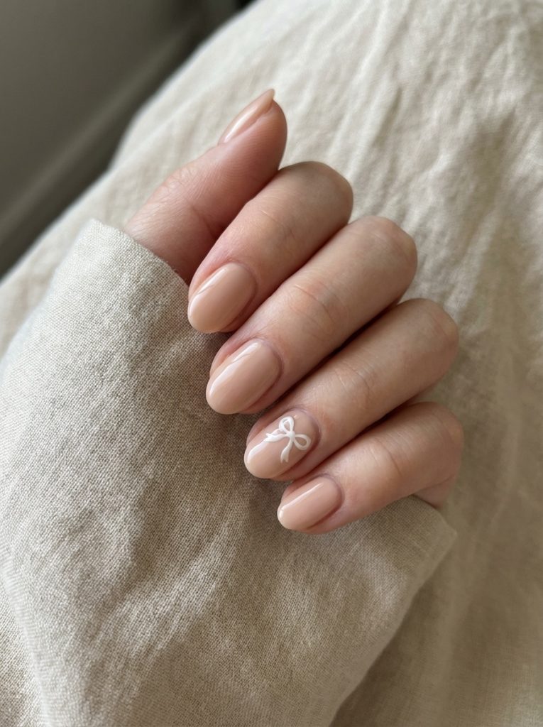 A relaxed hand wrapped in linen fabric displays nude wedding nails in a peachy beige polish, featuring a delicate white bow design on one accent nail.