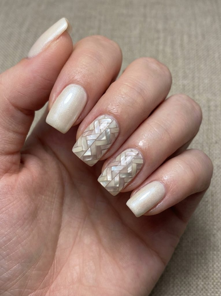 Natural hand posed against beige fabric showing pearl wedding nails in satin ivory polish and two nails decorated with mosaic-style woven patterns.