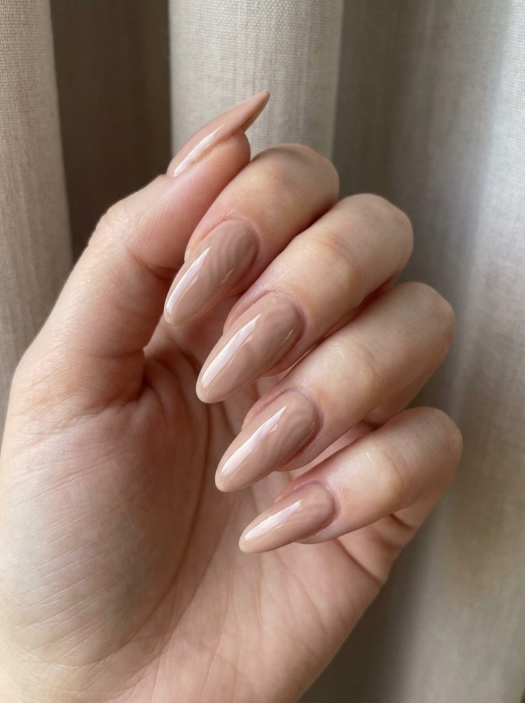 A close-up of a relaxed hand near soft fabric reveals nude wedding nails in a beige-peach tone, shaped into refined almond tips with a high-shine finish.
