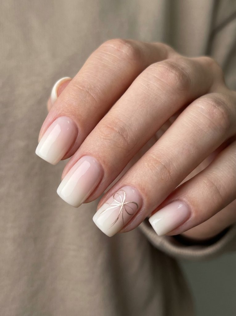 Natural hand pose showing glossy ombre wedding nails in pale pink fading to ivory white, highlighted by a minimalist bow accent on the ring finger.