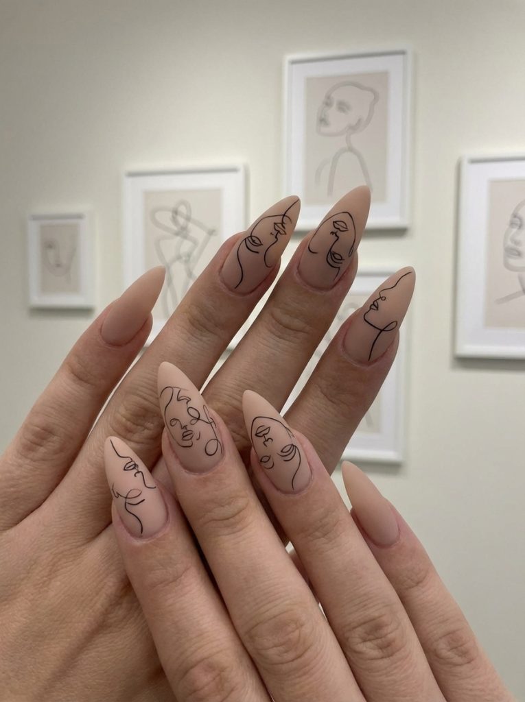 Close-up of almond-shaped matte wedding nails in blush nude with thin black line art portraits, posed against a wall with framed minimalist sketches.