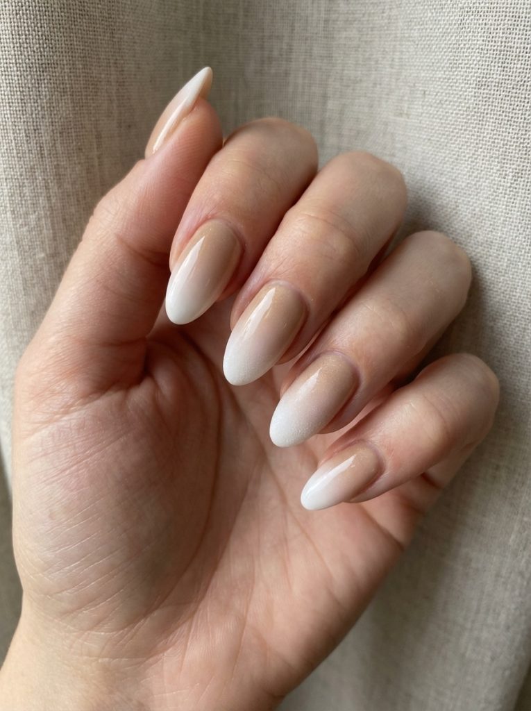 Hand resting near a textured linen curtain featuring almond ombre wedding nails that blend creamy nude into soft white with a luminous finish.