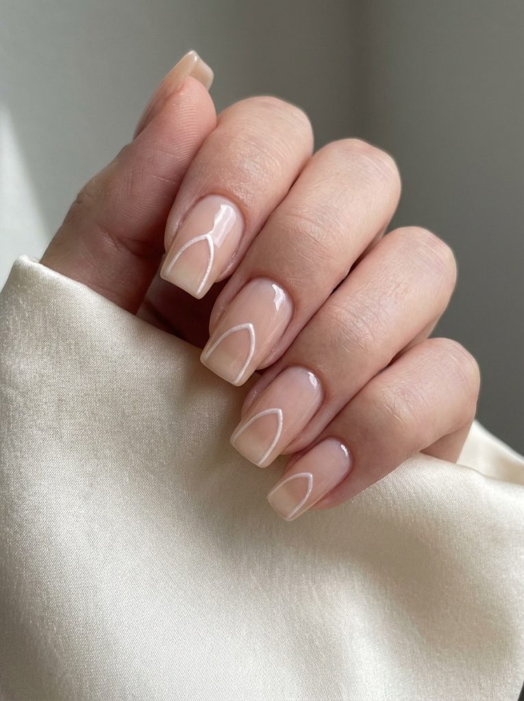 Hand posed against neutral background displaying nude wedding nails in peach-beige gloss with minimalist white curved French tip detailing on square-shaped nails.