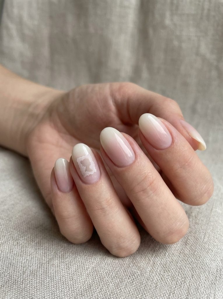 Rounded ombre wedding nails shown on a relaxed hand, blending blush into ivory and finished with a small decorative framed silhouette accent.