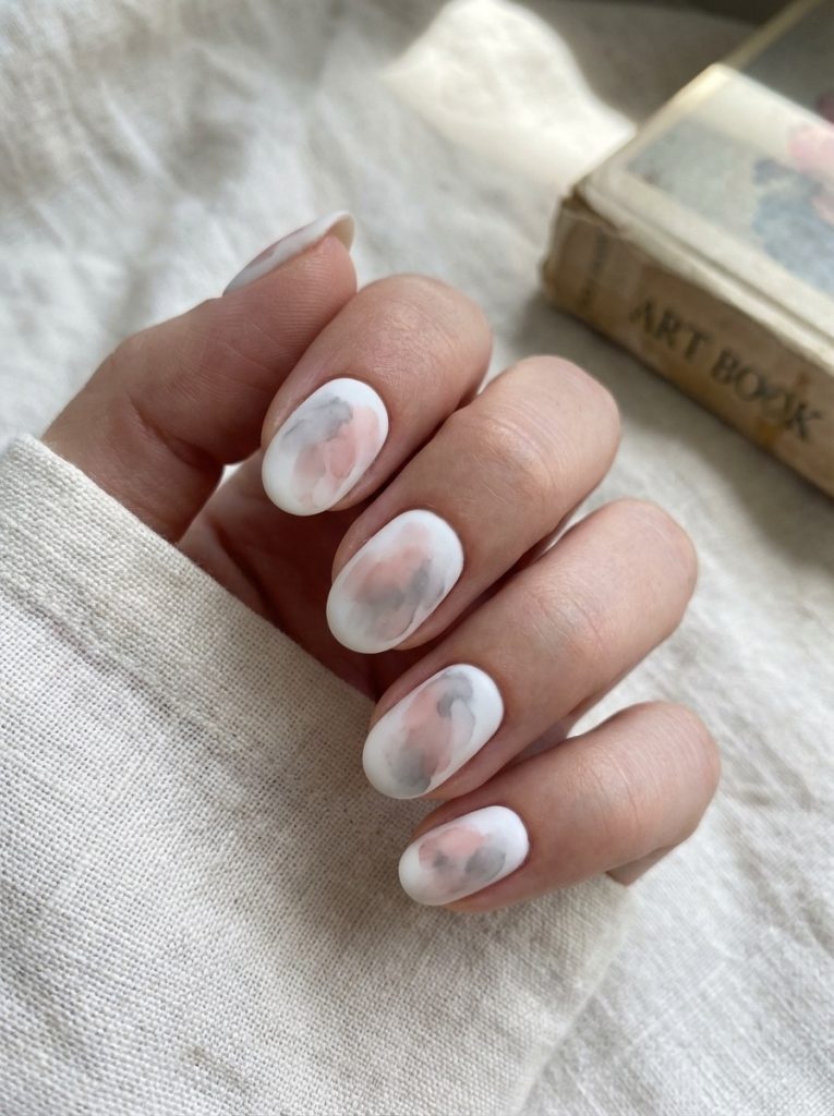 Close-up of a hand resting on textured fabric, featuring matte wedding nails in creamy white with subtle pink and gray watercolor floral details.