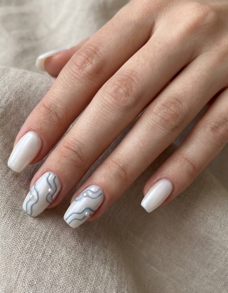Square pearl wedding nails photographed against beige fabric, featuring creamy white polish and flowing slate-blue lines accented by small pearl studs.