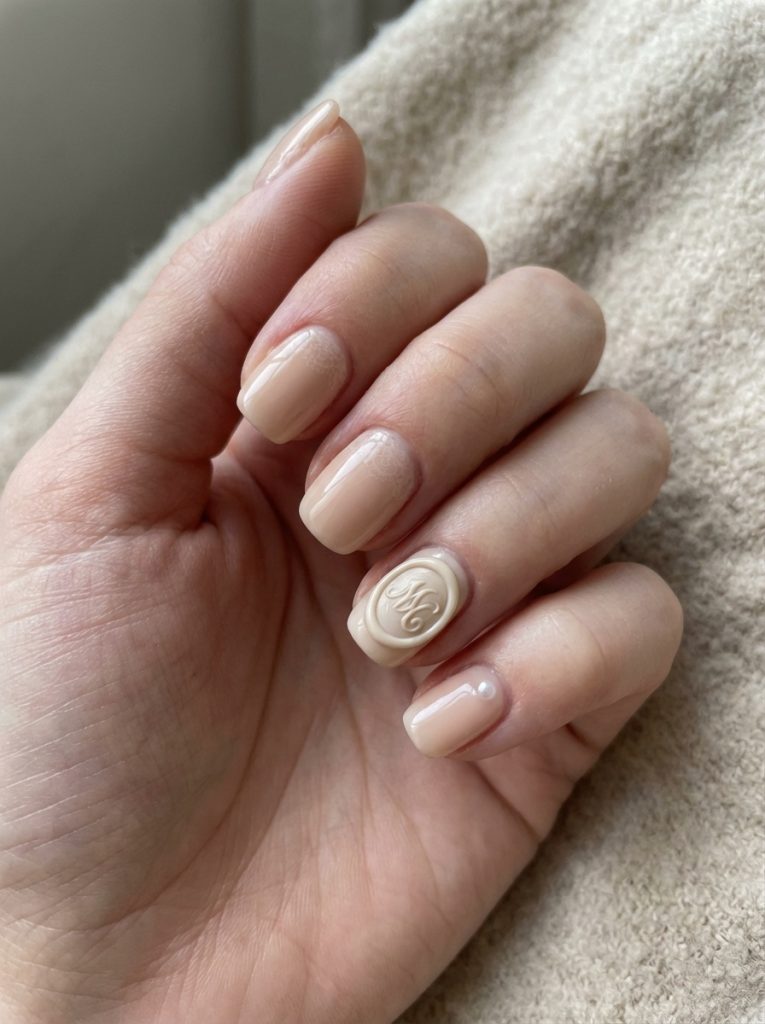Close-up of a hand displaying nude wedding nails in creamy beige polish with a raised cameo-style accent and a tiny pearl embellishment on short square nails.