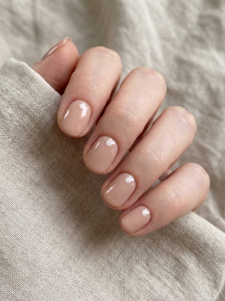 Close-up of hand resting on textured linen fabric showing nude wedding nails in creamy beige polish on short rounded nails with high-gloss finish.