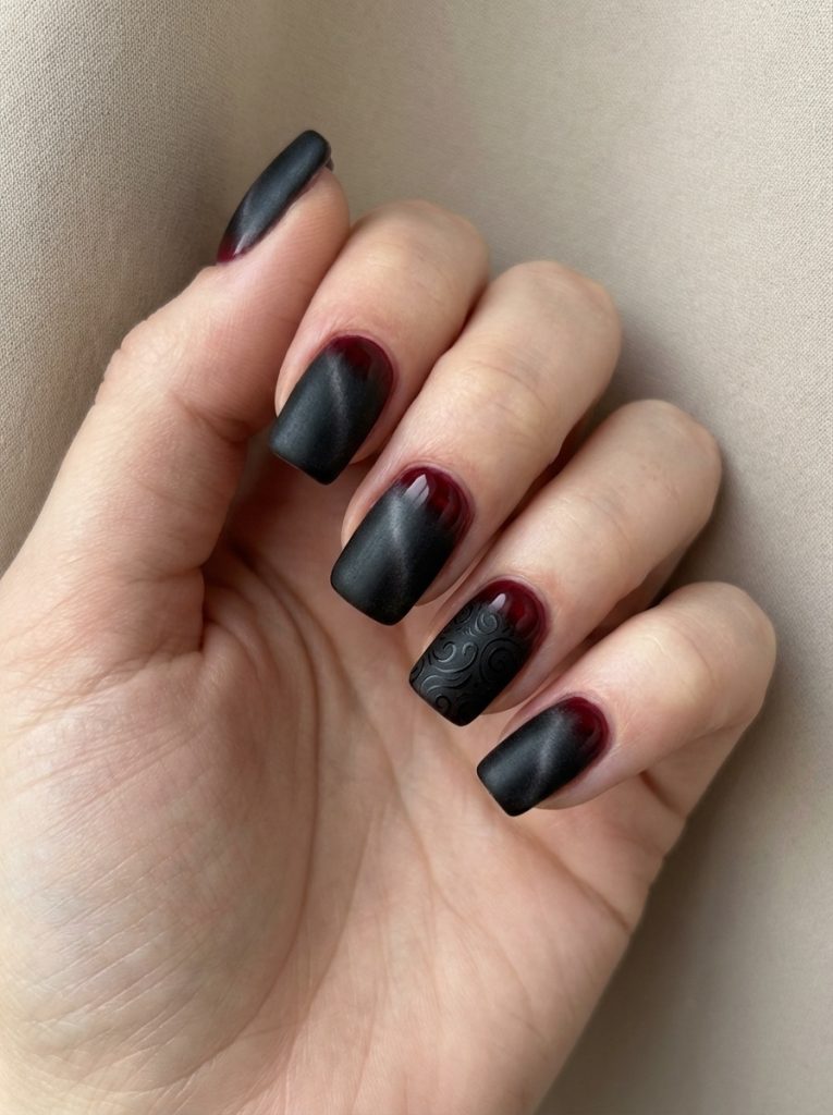 Hand resting against beige fabric displaying red and black wedding nails with a matte charcoal-to-burgundy ombré and one nail featuring raised swirl detailing.