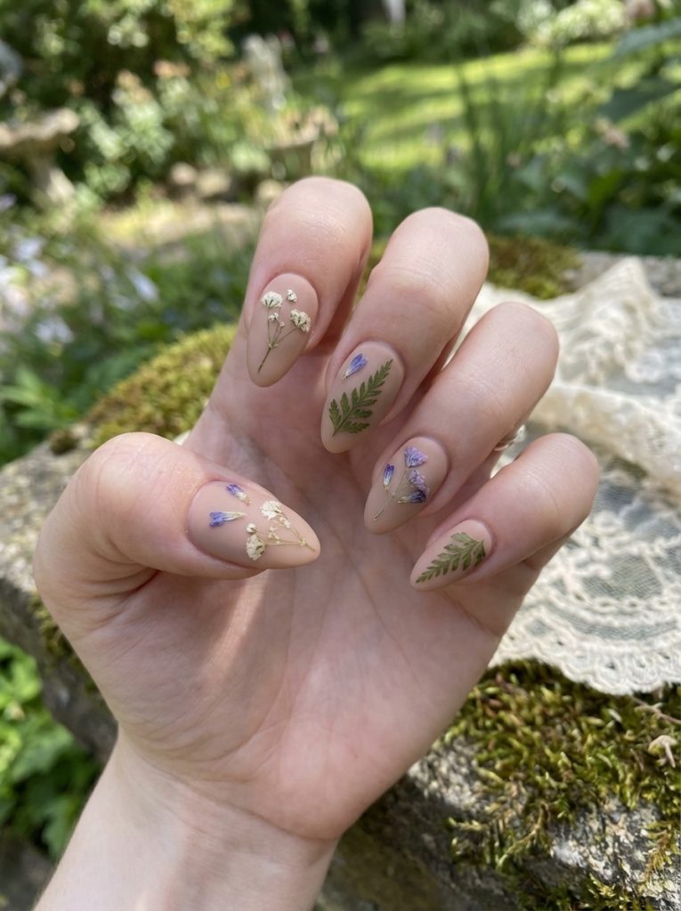Close-up of a hand resting on mossy stone featuring matte wedding nails in blush nude with miniature pressed florals and hand-painted green fern accents.