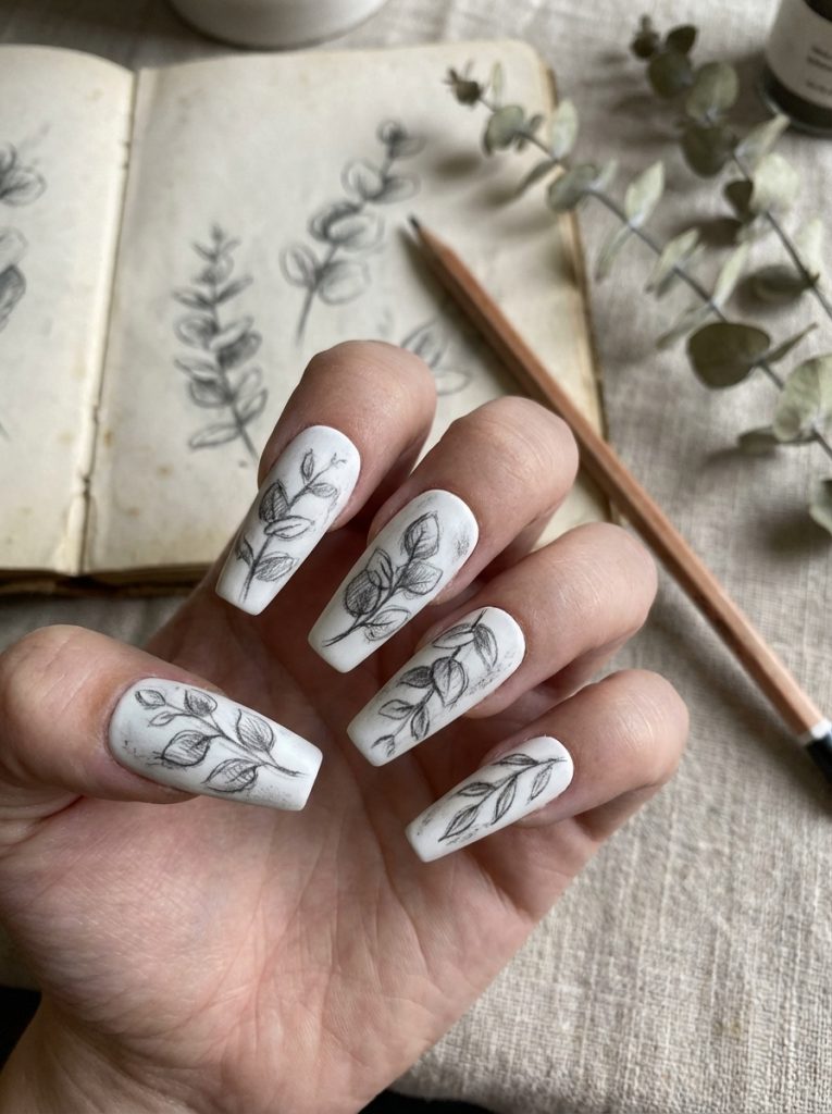 Hand holding matte wedding nails in creamy ivory with fine black leaf drawings, long coffin-shaped tips, styled over linen fabric beside an open sketchbook and pencil.