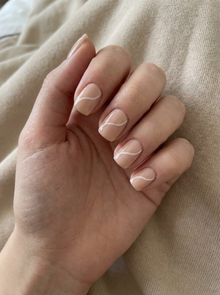 A close-up of a hand resting on soft beige fabric features nude wedding nails with a translucent beige base, modern white wave accents, and short square shaping.