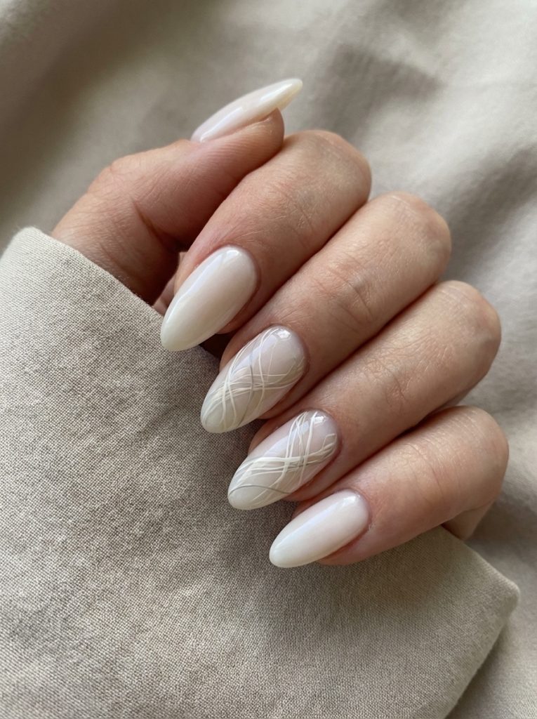 Hand resting against beige fabric showing pearl wedding nails in creamy off-white polish with intricate white swirl art on the middle fingers.